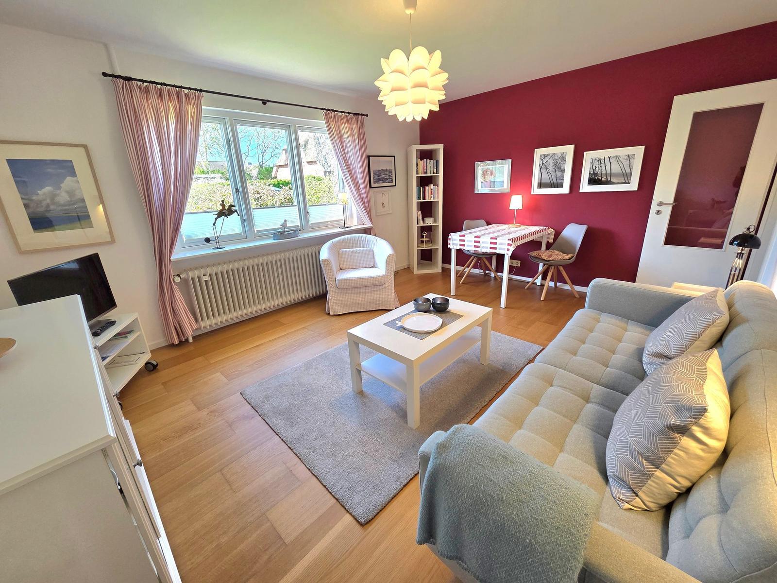 Wohn- und Essbereich mit Sofa, Tisch und Fenster. Rote Wand und Holzboden.