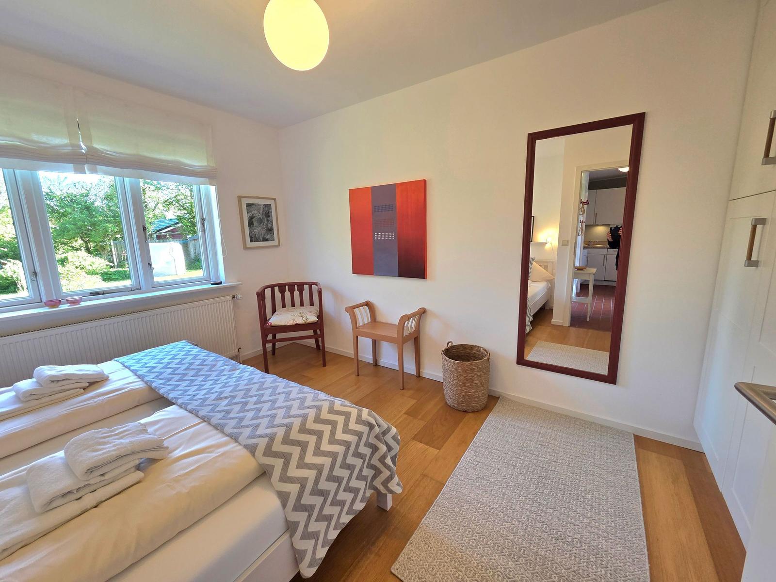 Schlafzimmer mit Bett, Fenster, zwei Stühlen und Spiegel. Holzboden und Wandbild.
