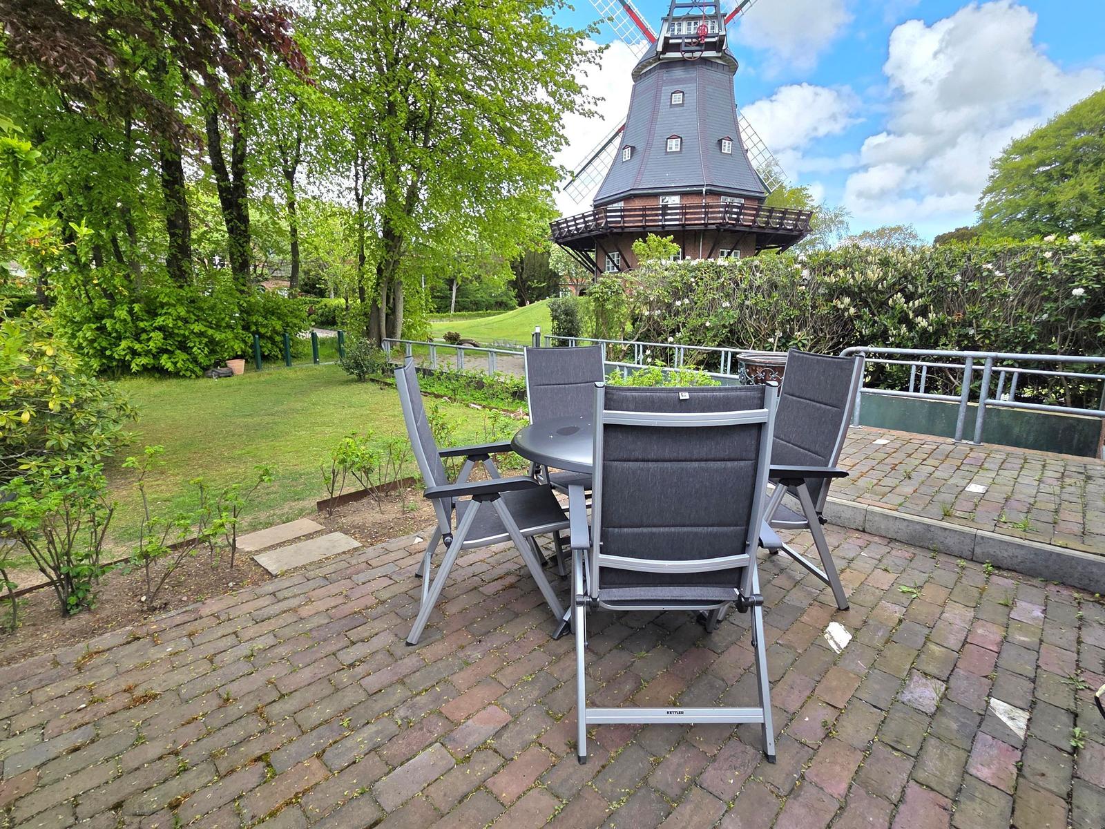 Terrasse mit Tisch und Stühlen, Blick auf Windmühle und Grünanlagen.