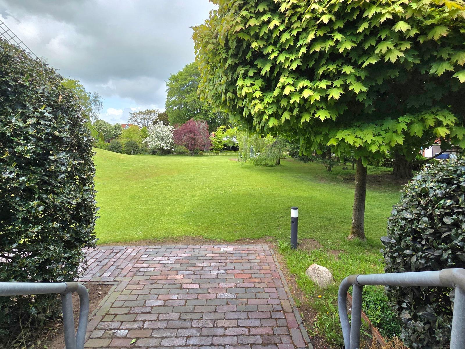 Pflasterweg führt durch grünen Garten mit Bäumen und Sträuchern.