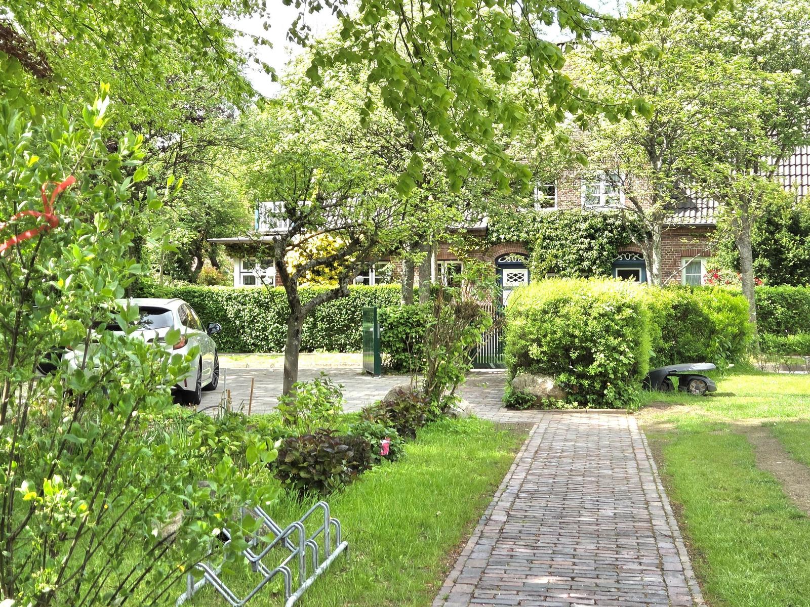 Ein gepflasterter Weg führt zu einem Haus mit Garten und Bäumen. Ein Auto steht auf dem Hof.