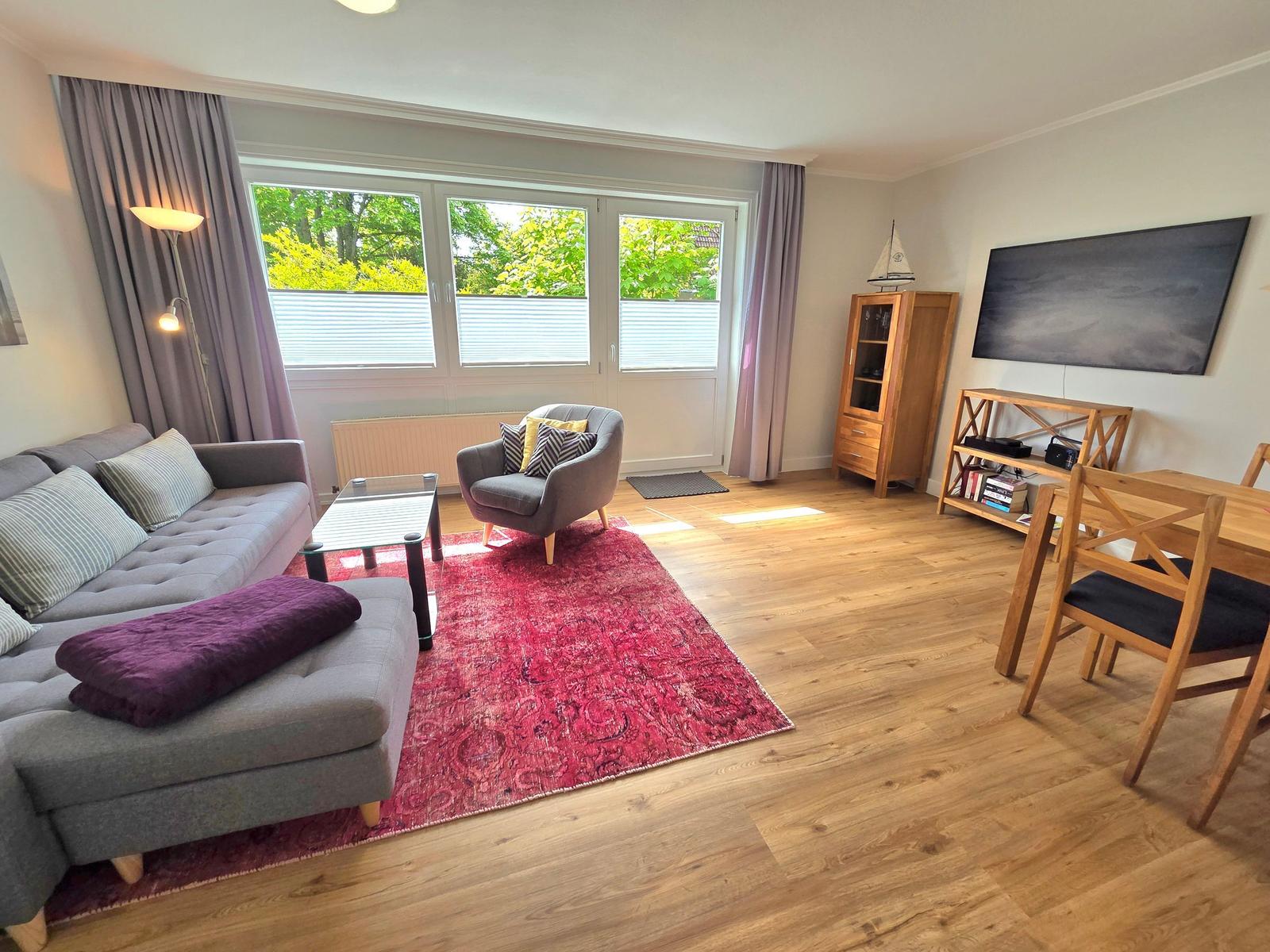Wohnzimmer mit Sofa, Sessel, Esstisch und TV. Holzboden und großes Fenster.