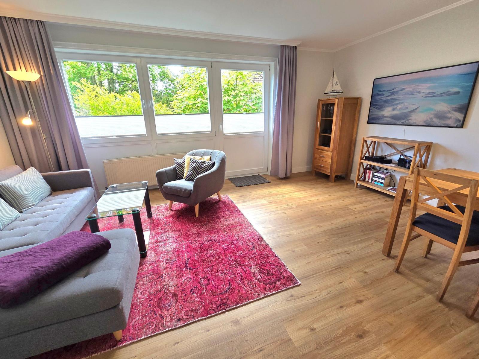 Wohnzimmer mit Sofa, Sessel, Teppich und Fenster zur grünen Aussicht.