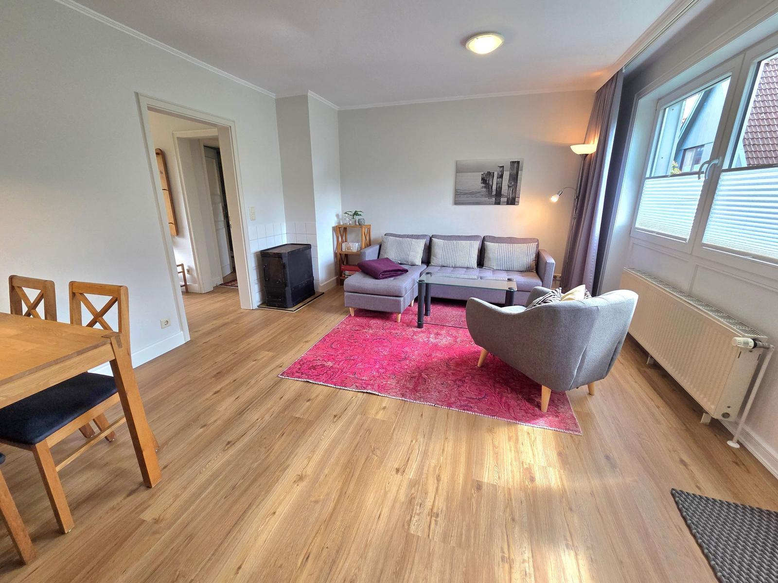 Wohnzimmer mit Sofa, Sessel, Esstisch und Holzboden. Fenster mit Jalousien.