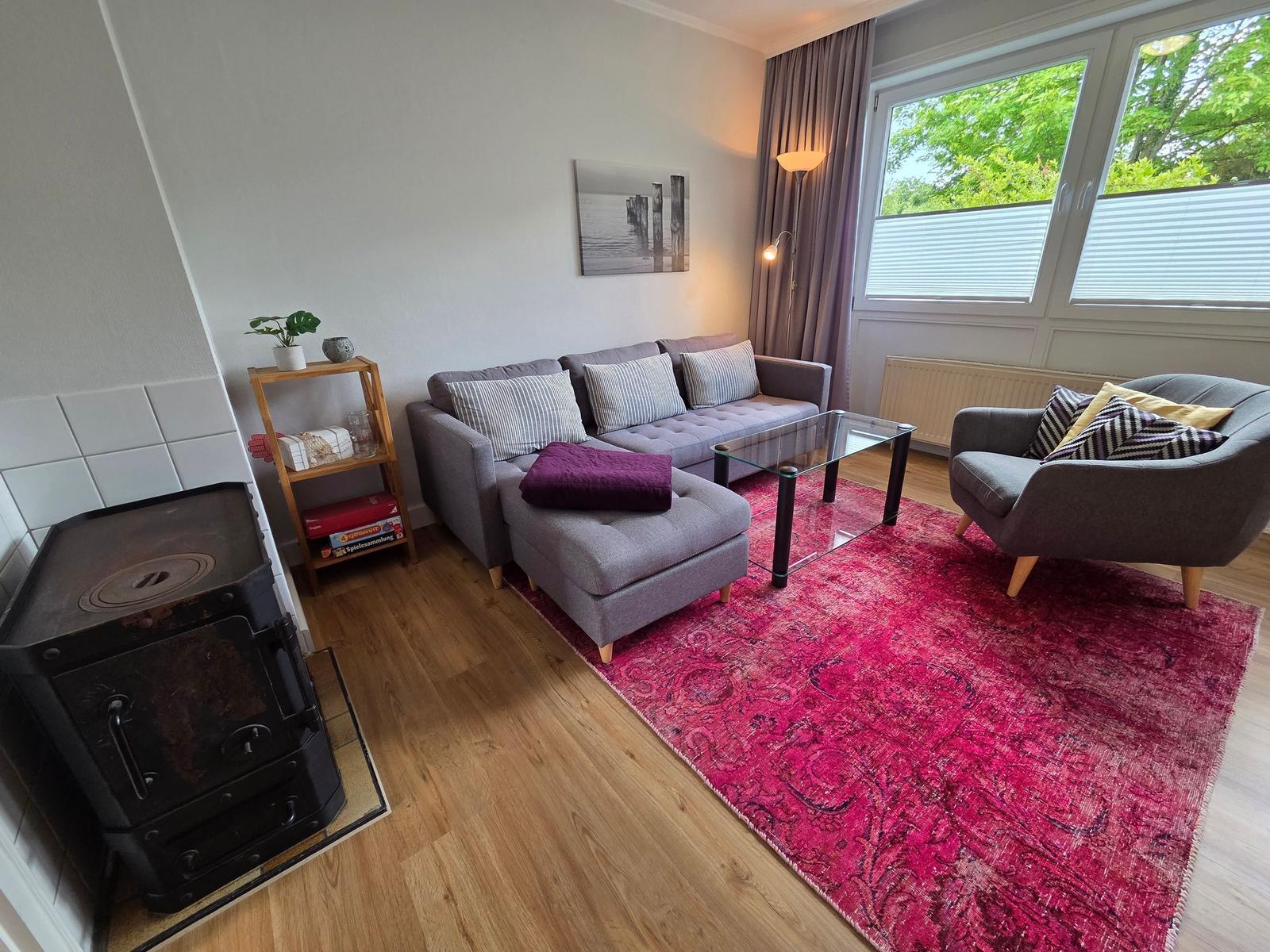 Wohnzimmer mit grauem Sofa, rotem Teppich und Glas-Tisch. Fenster mit Blick auf Grün.