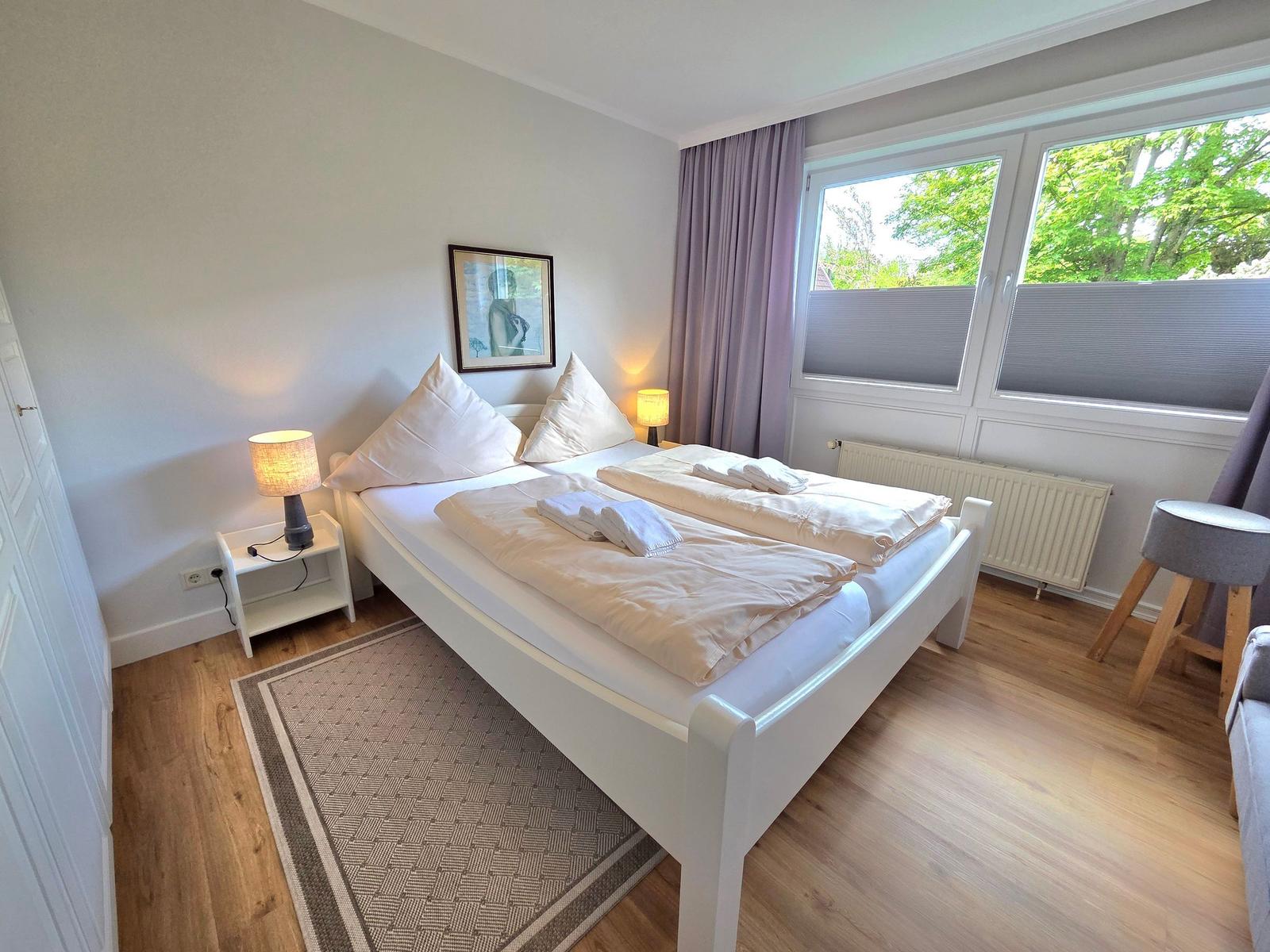 Schlafzimmer mit Doppelbett, Nachttisch und Fenster mit Blick auf Bäume.