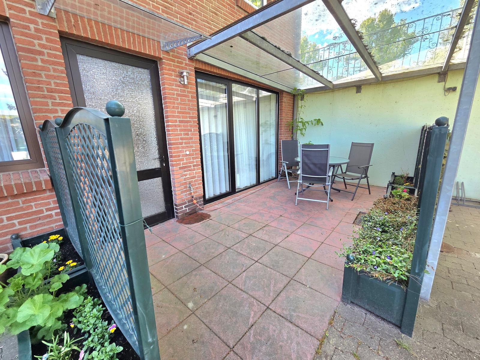 Terrasse mit Sitzgruppe unter Glasdach, umgeben von Pflanzen und Zäunen.