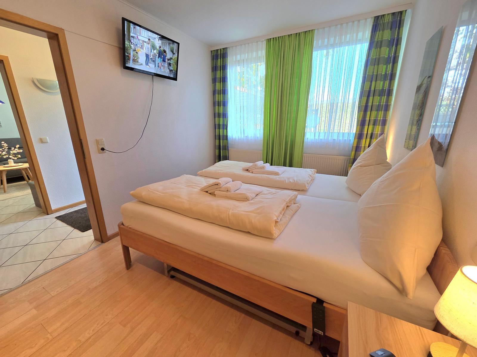 Zimmer mit Doppelbett, TV und Fenster mit grünen Vorhängen.