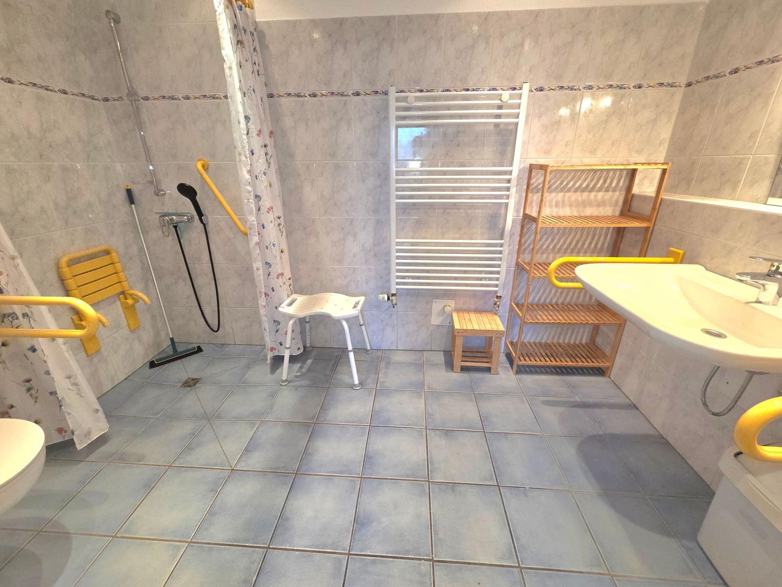 Badezimmer mit Dusche, WC, Waschbecken und Pflegehilfsmitteln.