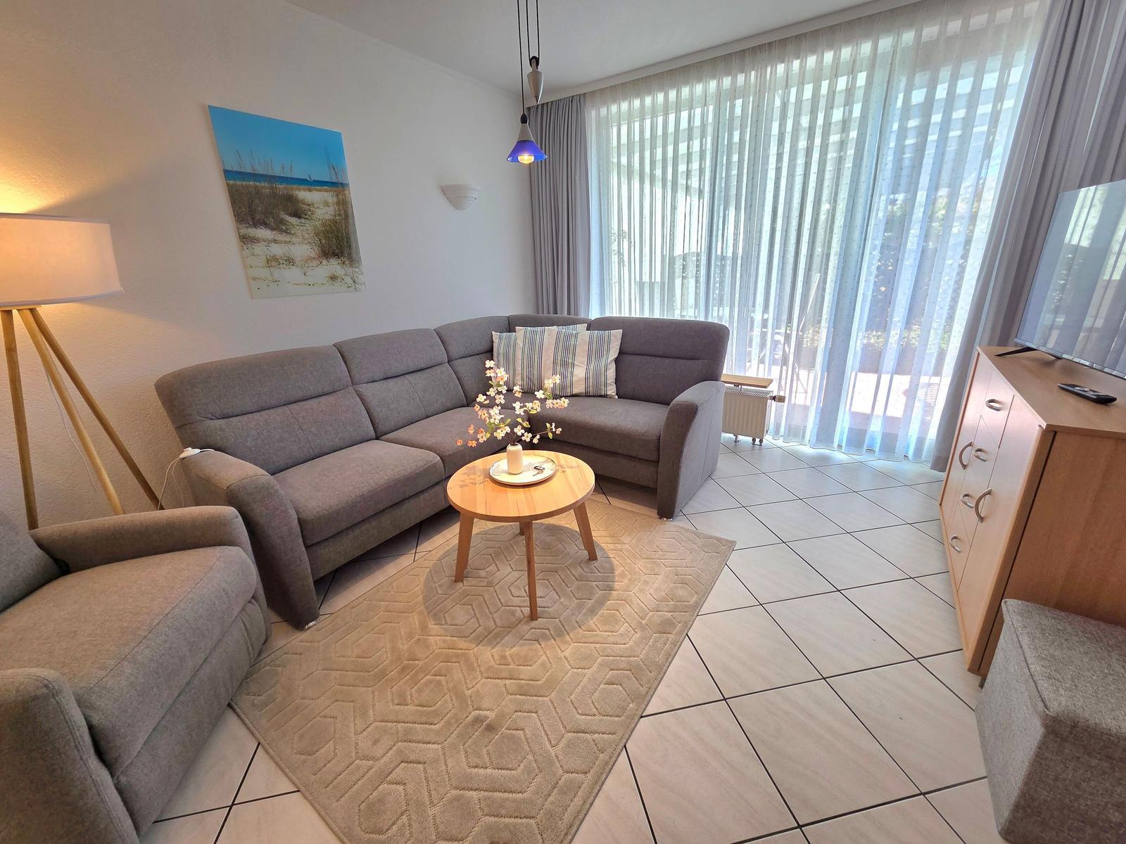 Wohnzimmer mit grauem Sofa, Holztisch und TV-Schrank. Große Fenster mit Vorhängen.