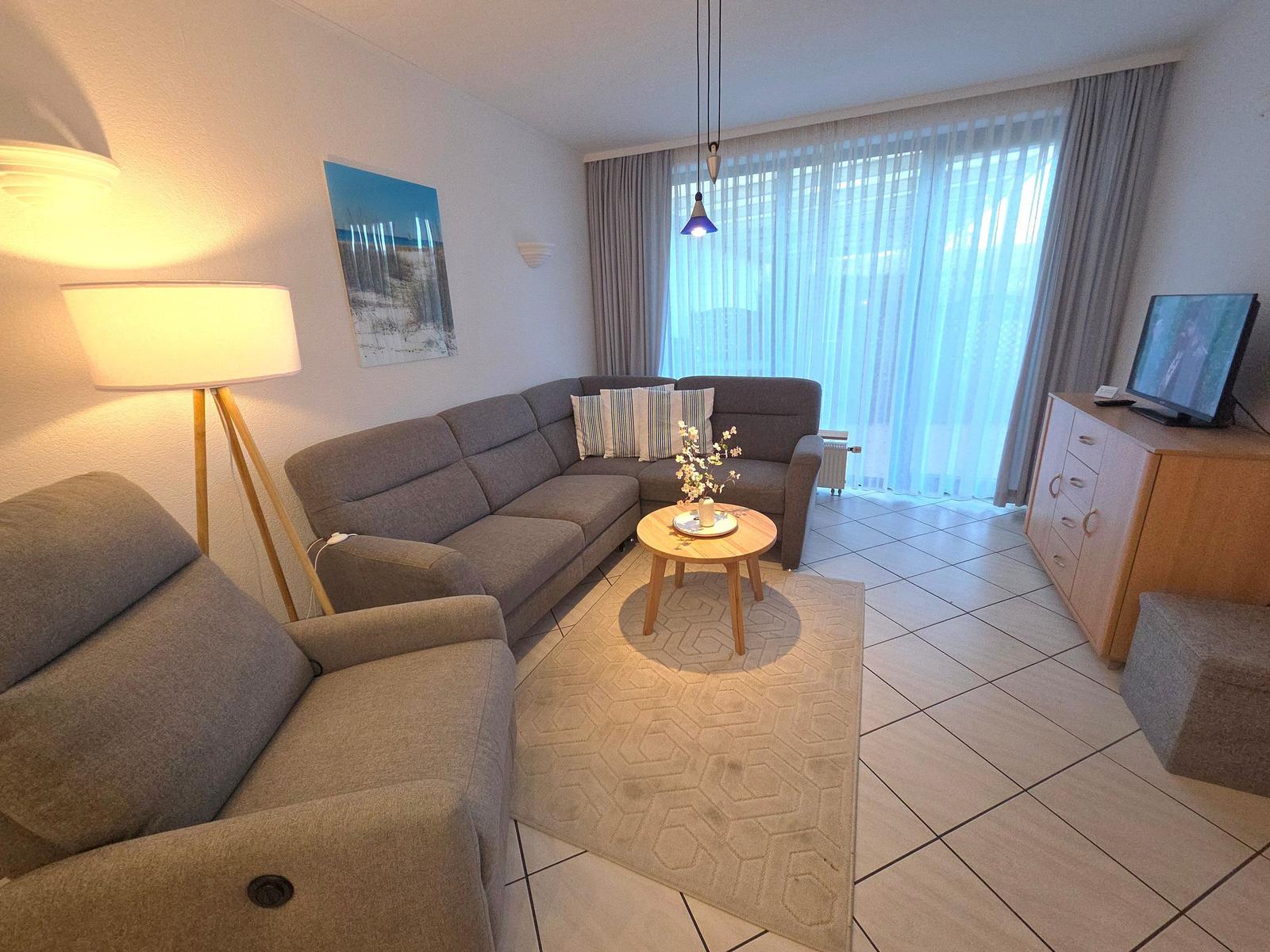 Wohnzimmer mit grauem Sofa, Beistelltisch und TV. Fenster mit Vorhängen.