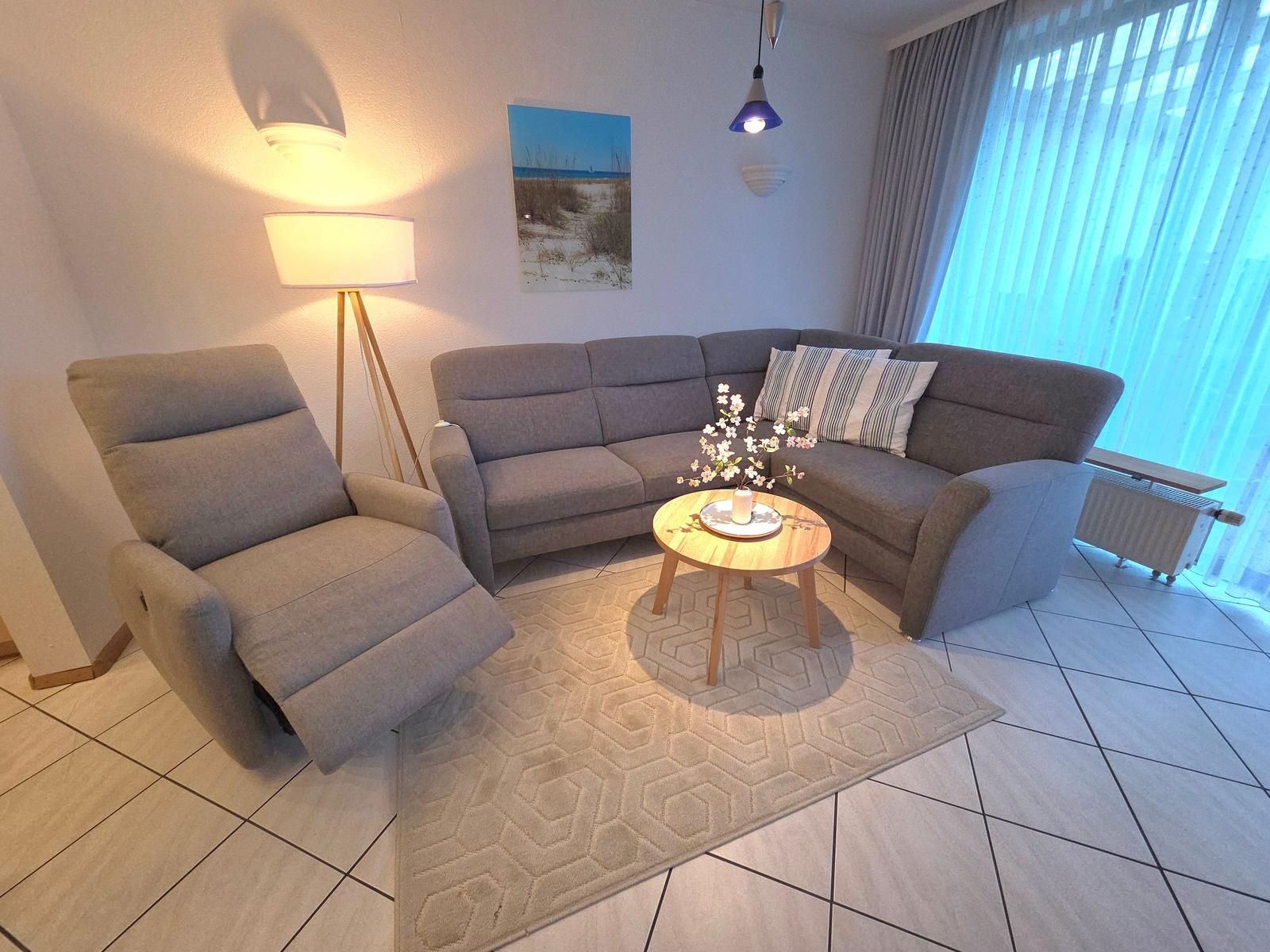 Wohnzimmer mit grauem Sofa, Sessel, Tisch und Teppich. Fenster mit Jalousie und Wandbild.