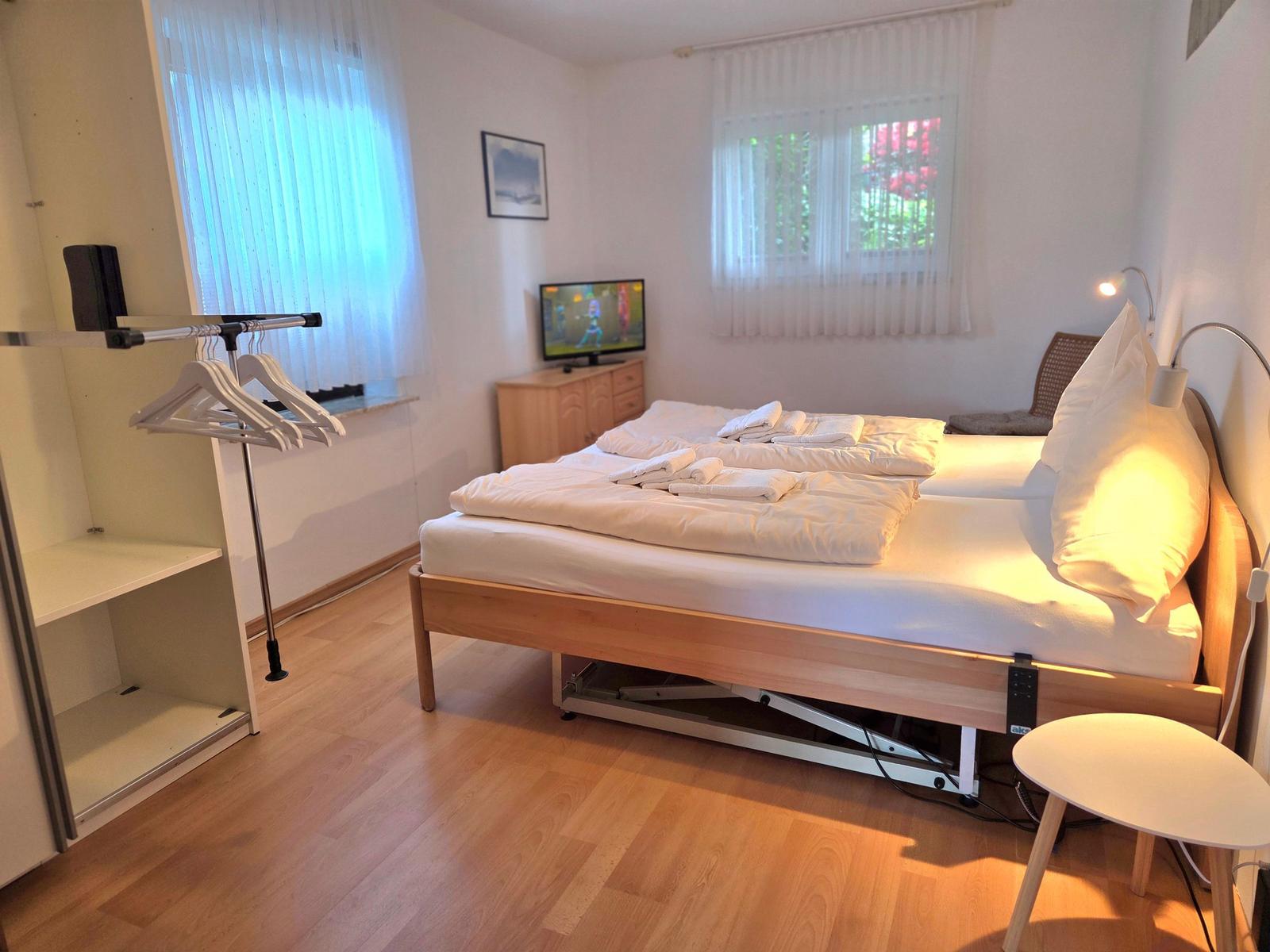 Bedroom with double bed, clothes rack, and TV. Wooden floor and windows with blinds.