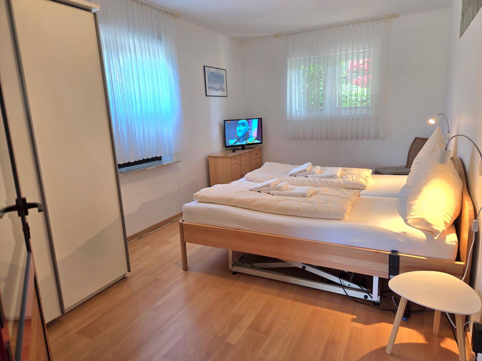 Bedroom with bed, TV, and wooden floor. Windows with white curtains.