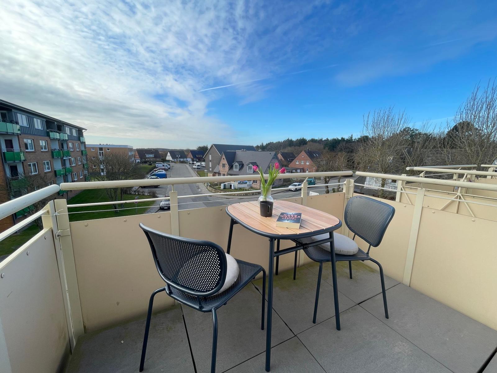 Balkon mit Tisch und zwei Stühlen. Blick auf Straße und Wohnhäuser.