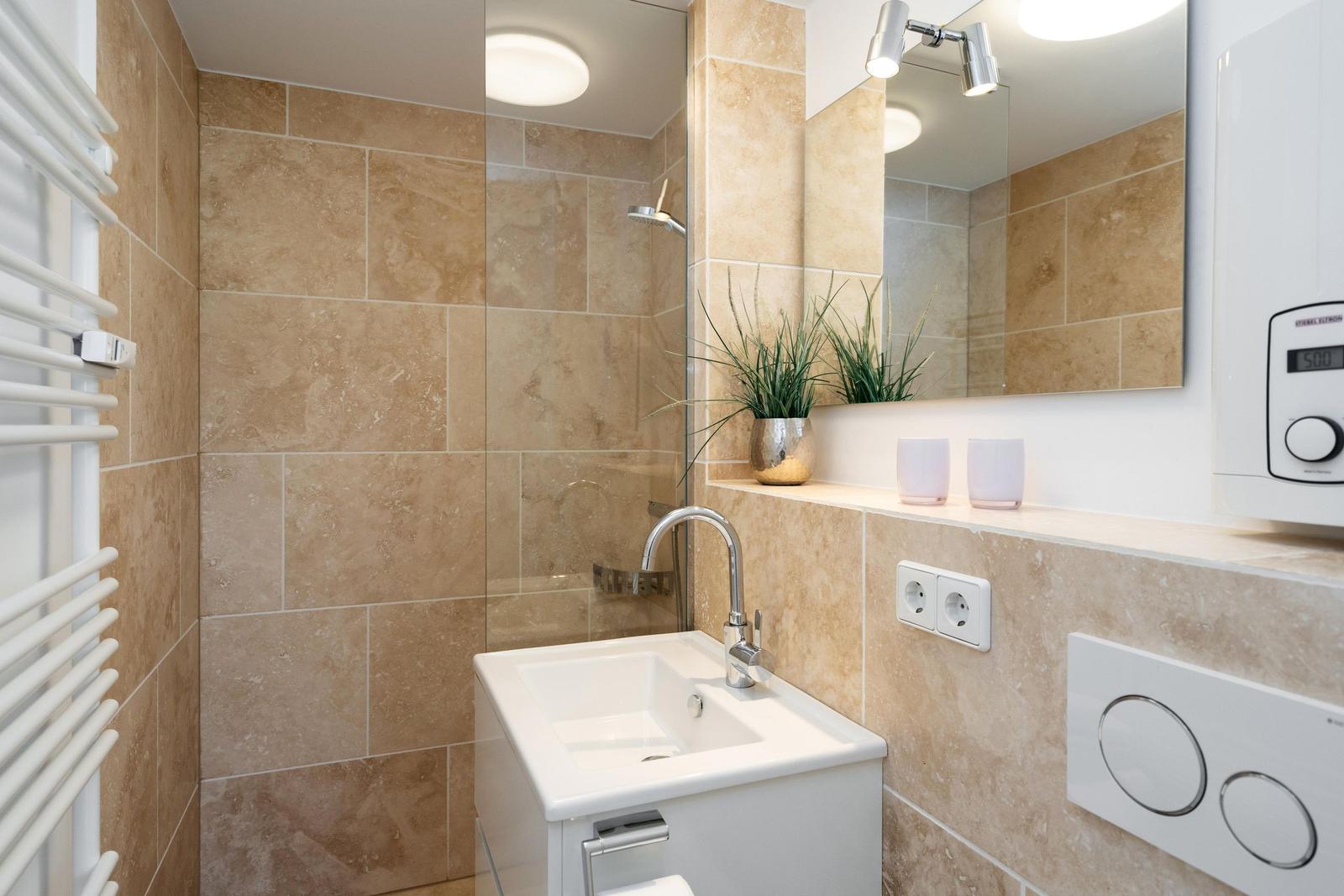 Bad mit Dusche, Waschbecken und Spiegel. Beleuchtung und Pflanze sind sichtbar.