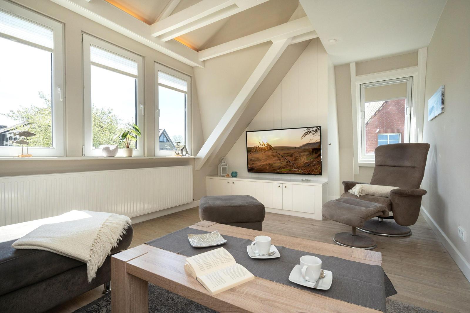 Wohnzimmer mit Fernseher, Sessel und Tisch. Drei Fenster und schräge Decke.