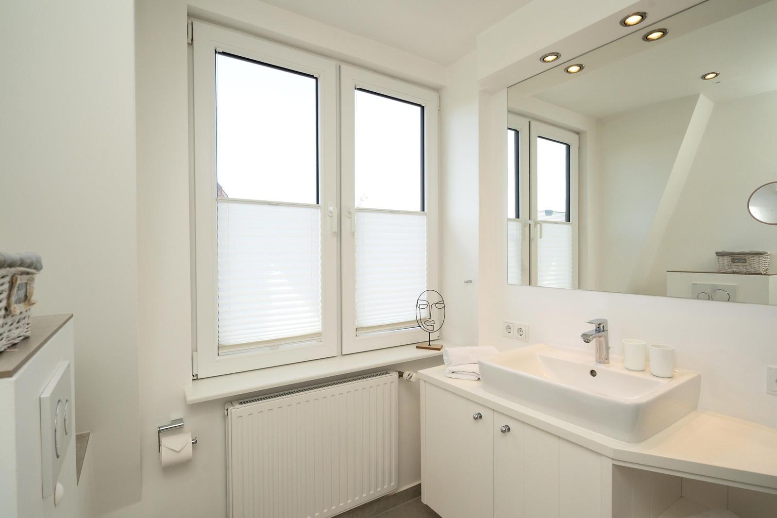 Badezimmer mit Fenster, Spiegel und Waschtisch. Weiße Einrichtung und Heizung.