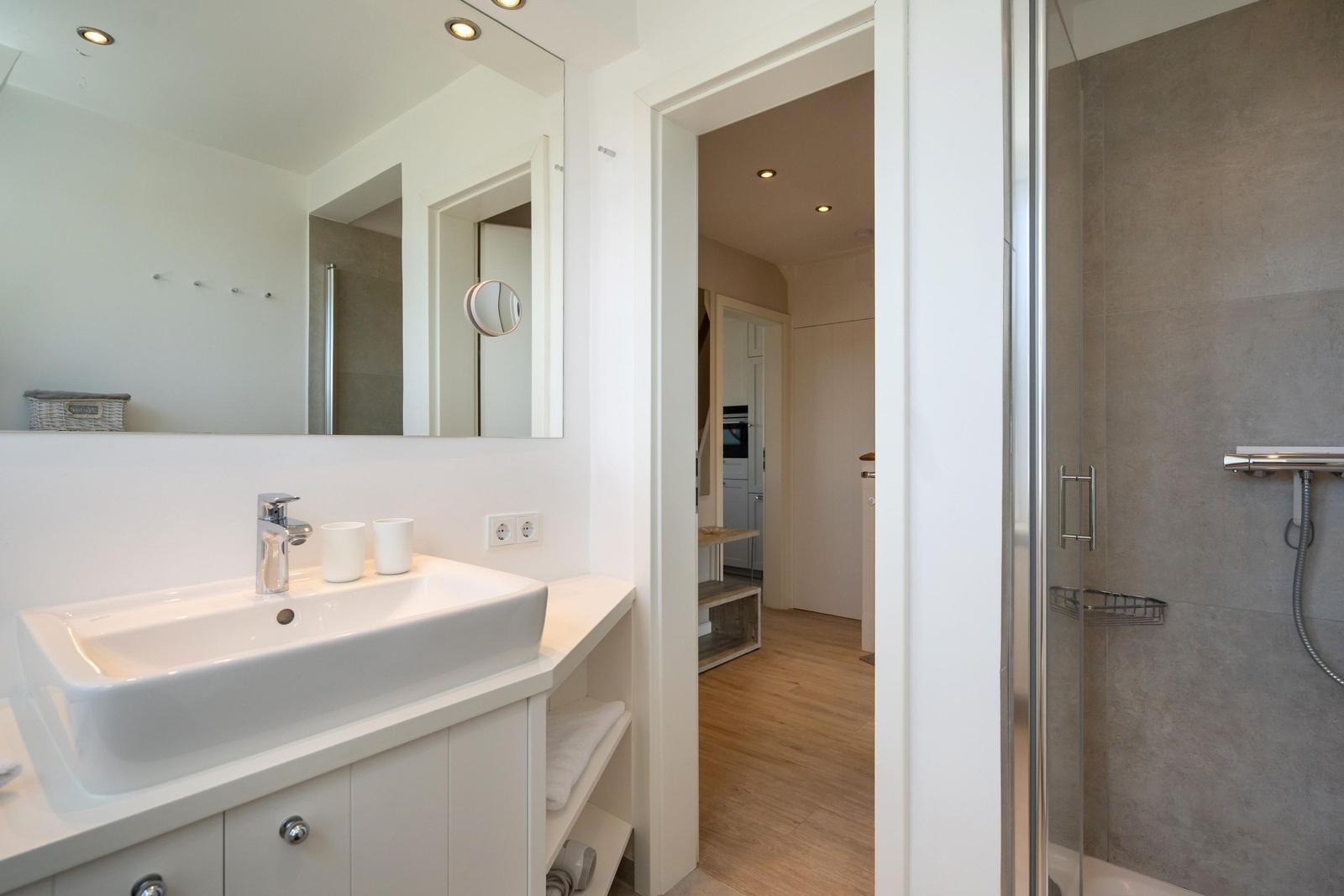 Badezimmer mit Waschbecken, Spiegel und Dusche. Blick in angrenzenden Raum.