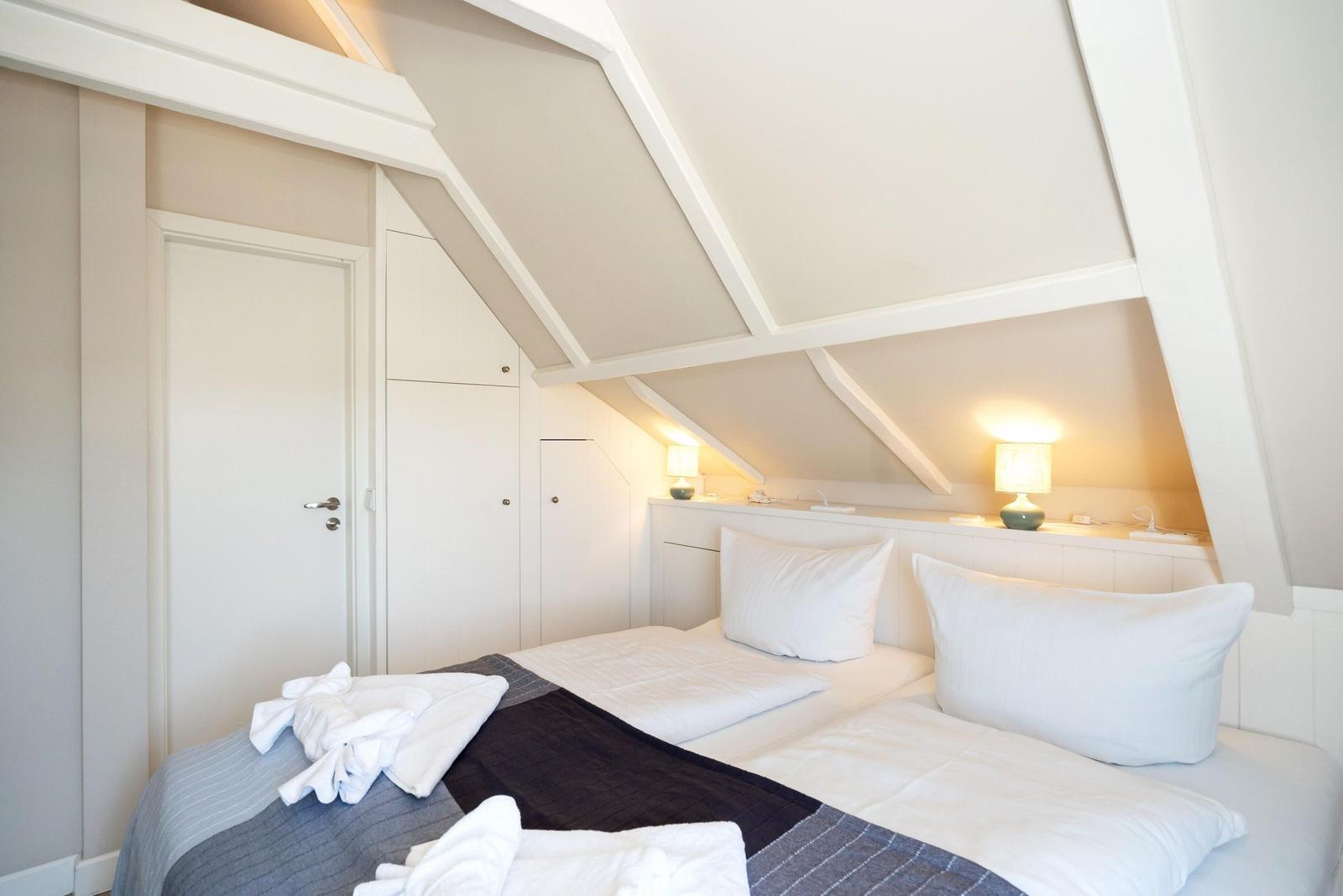 Schlafzimmer mit Dachschrägen, Bett, zwei Nachttische mit Lampen und Tür.