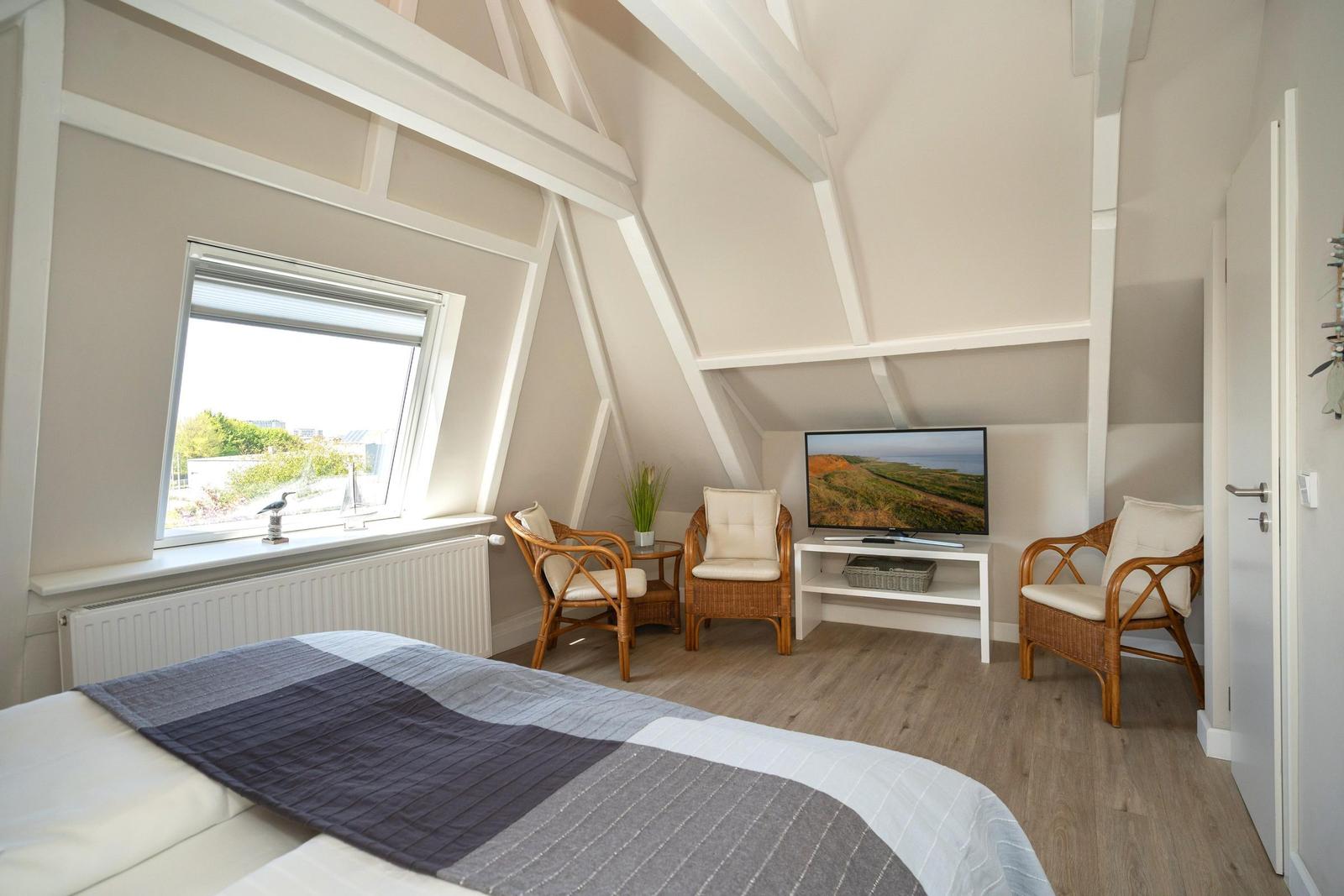 Schlafzimmer mit Dachschräge, Bett, Fernseher und drei Stühlen. Fenster mit Blick nach draußen.