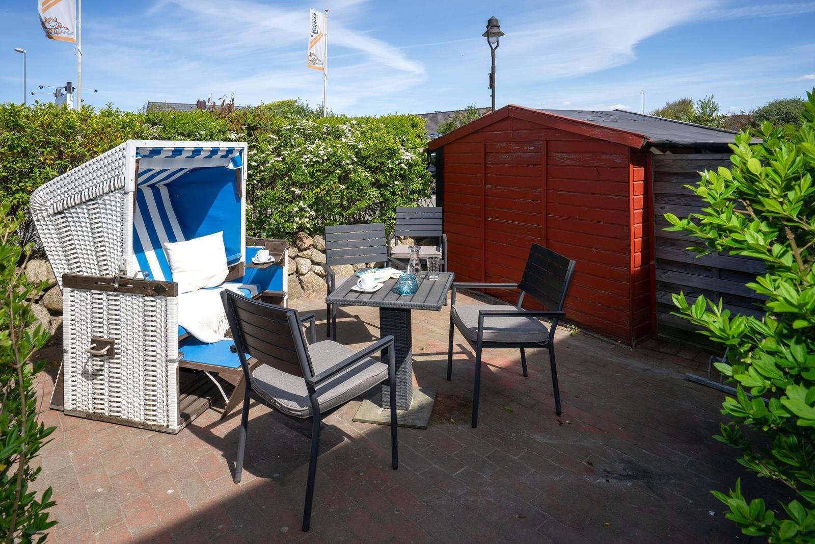 Terrasse mit Strandkorb, Tisch und Stühlen neben rotem Holzschuppen.
