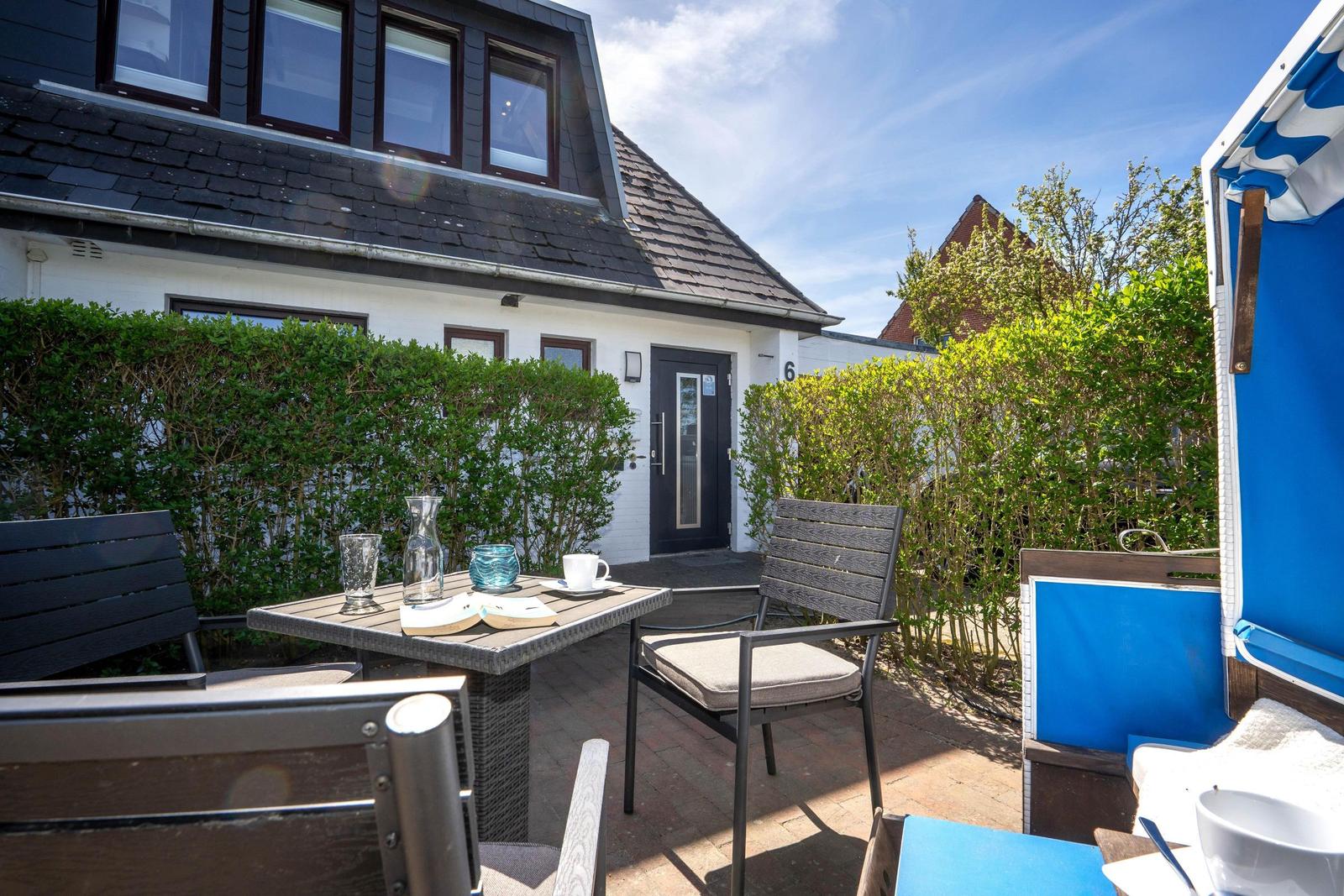Terrasse mit Tisch, Stühlen und Strandkorb. Hinterhof mit Hecke und Haus.