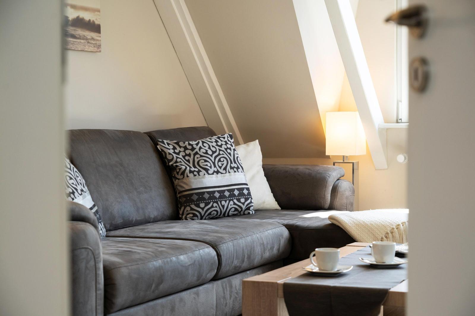 Ein grauer Sofa mit Kissen unter schrägen Dachbalken. Tisch mit zwei Tassen.