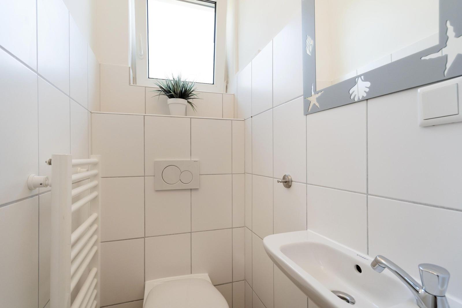 Badezimmer mit Toilette, Waschbecken und Fenster. Weiße Fliesen und Spiegel mit Meeresmotiv.