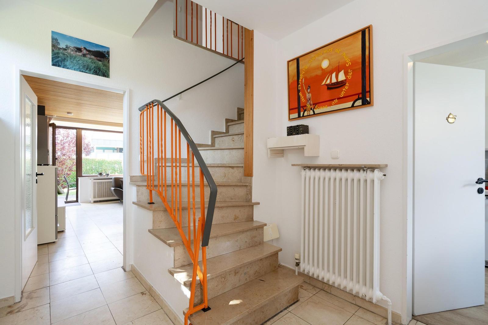 Treppe mit orangefarbenem Geländer führt nach oben. Wandbilder und Heizkörper sind sichtbar.