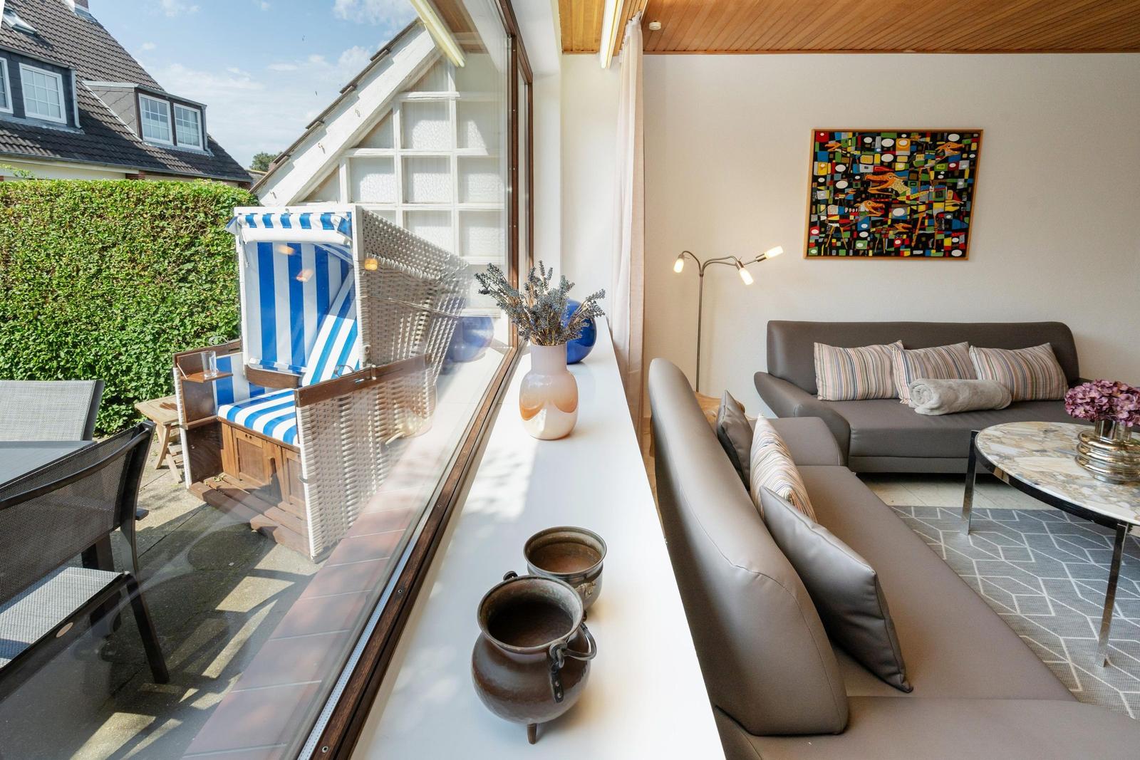 Ein Wohnzimmer mit Sofa, Tisch und Wandbild. Durch Fenster ist eine Terrasse mit Strandkorb sichtbar.