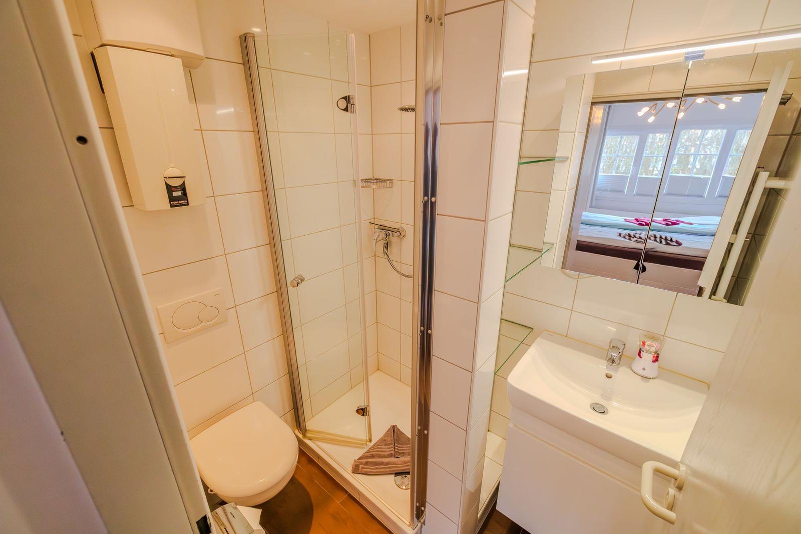Badezimmer mit Dusche, Waschbecken und Toilette. Spiegel reflektiert das Fenster.