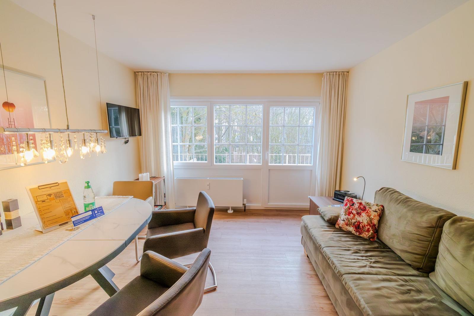 Wohn- und Essbereich mit Sofa, Tisch und Fenster. Wandbild und Hängelampe sichtbar.