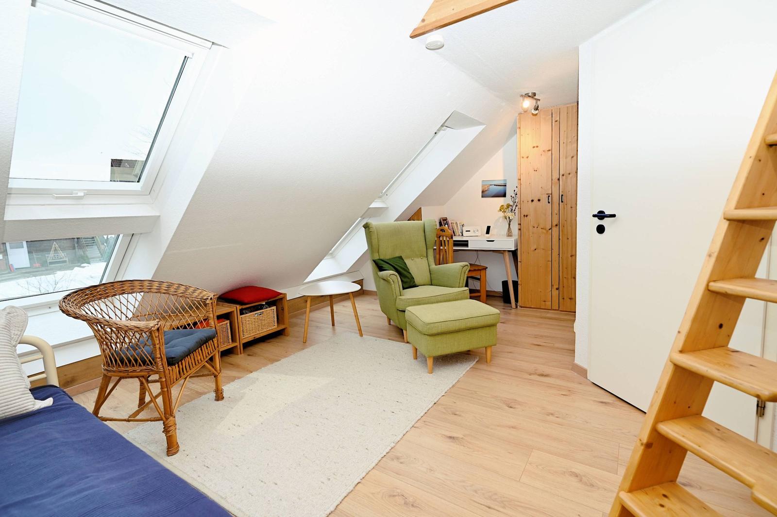 Ein Schlafzimmer mit Dachschrägen, Holzboden und einem grünen Sessel. Links ein Bett, rechts eine Treppe.