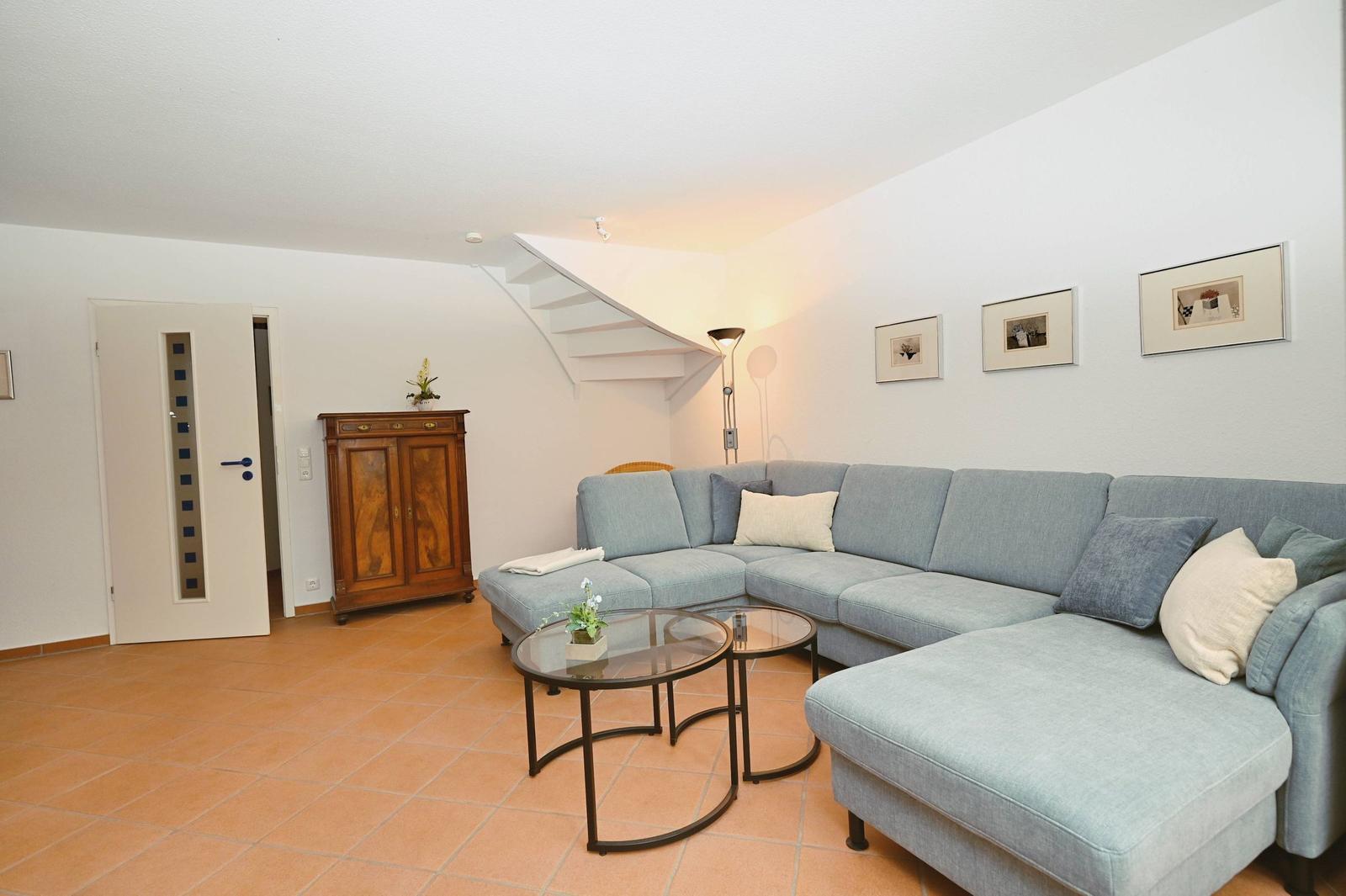 Wohnzimmer mit Ecksofa, Glas-Tisch und Holzschrank. Treppenhaus und Wandbilder sichtbar.