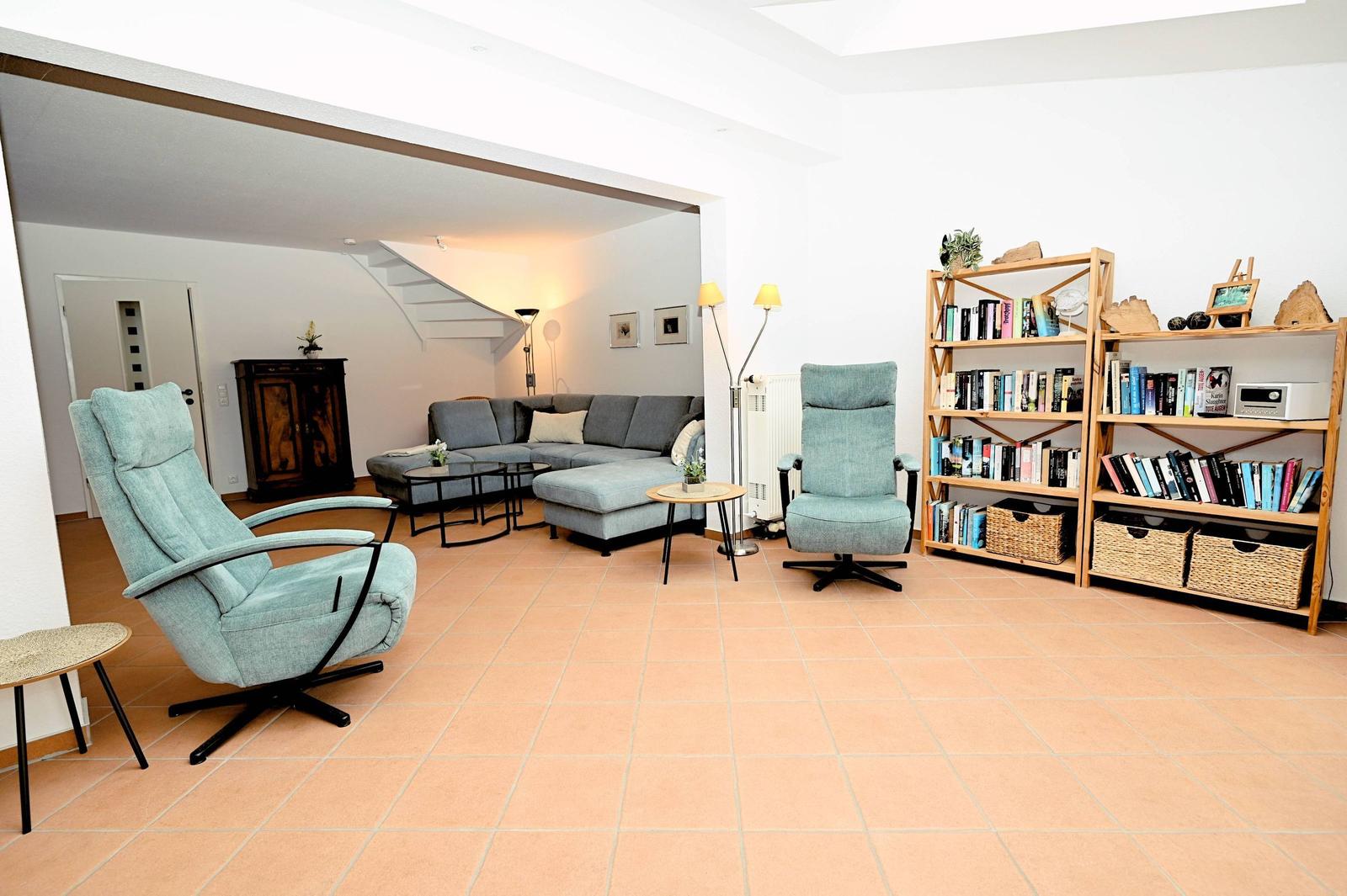 Wohnzimmer mit Sofa, zwei Sesseln und Bücherregal. Terrakotta-Boden und helle Wände.