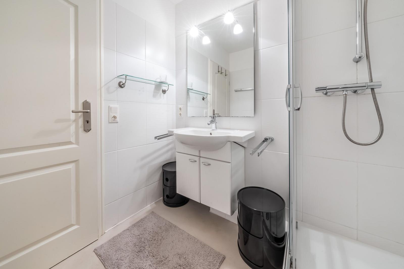 Bathroom with shower, sink, and mirror. White tiles and black trash cans.