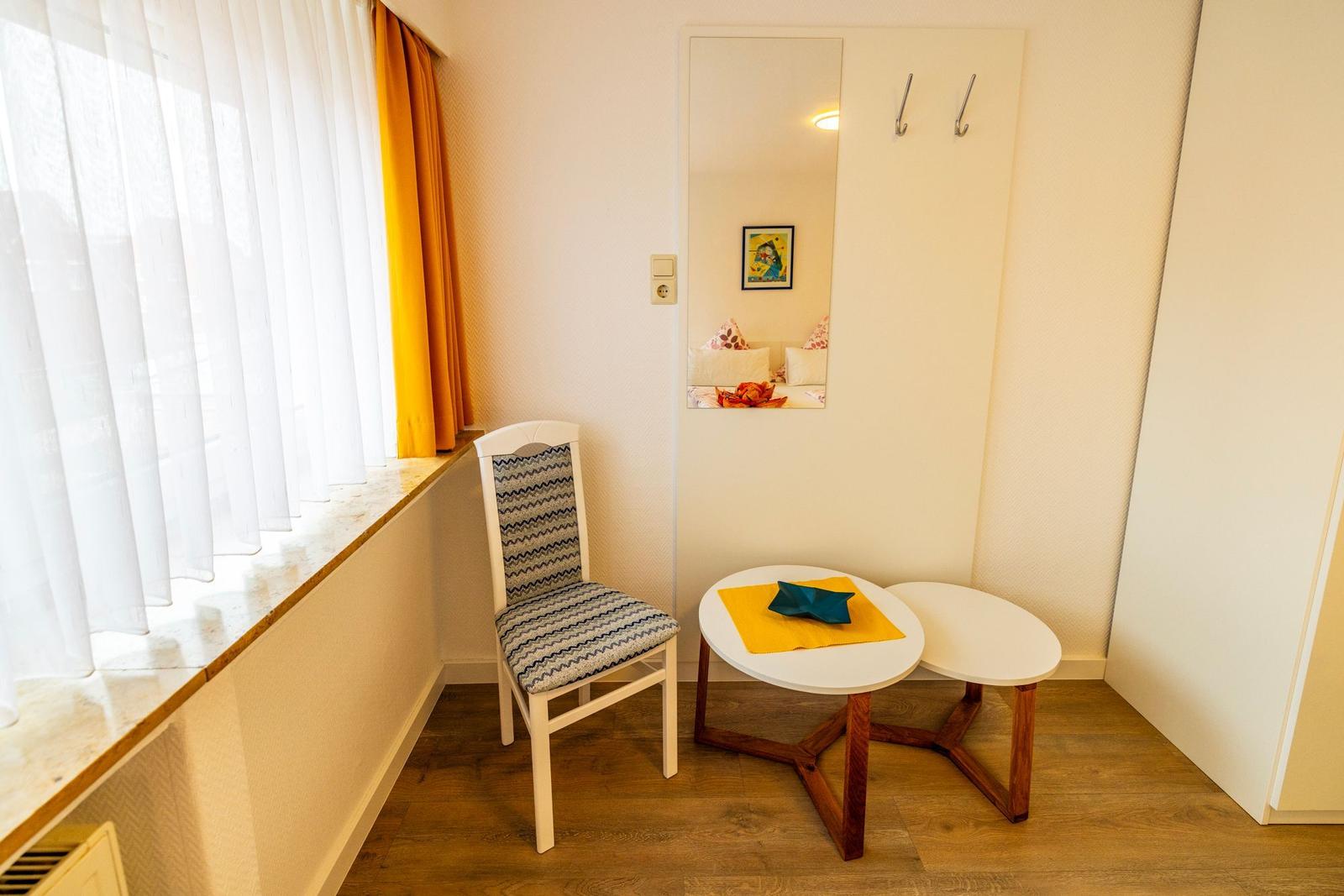 Ein Raum mit Stuhl, Tisch und Spiegel an der Wand. Fenster mit weißen und gelben Vorhängen.