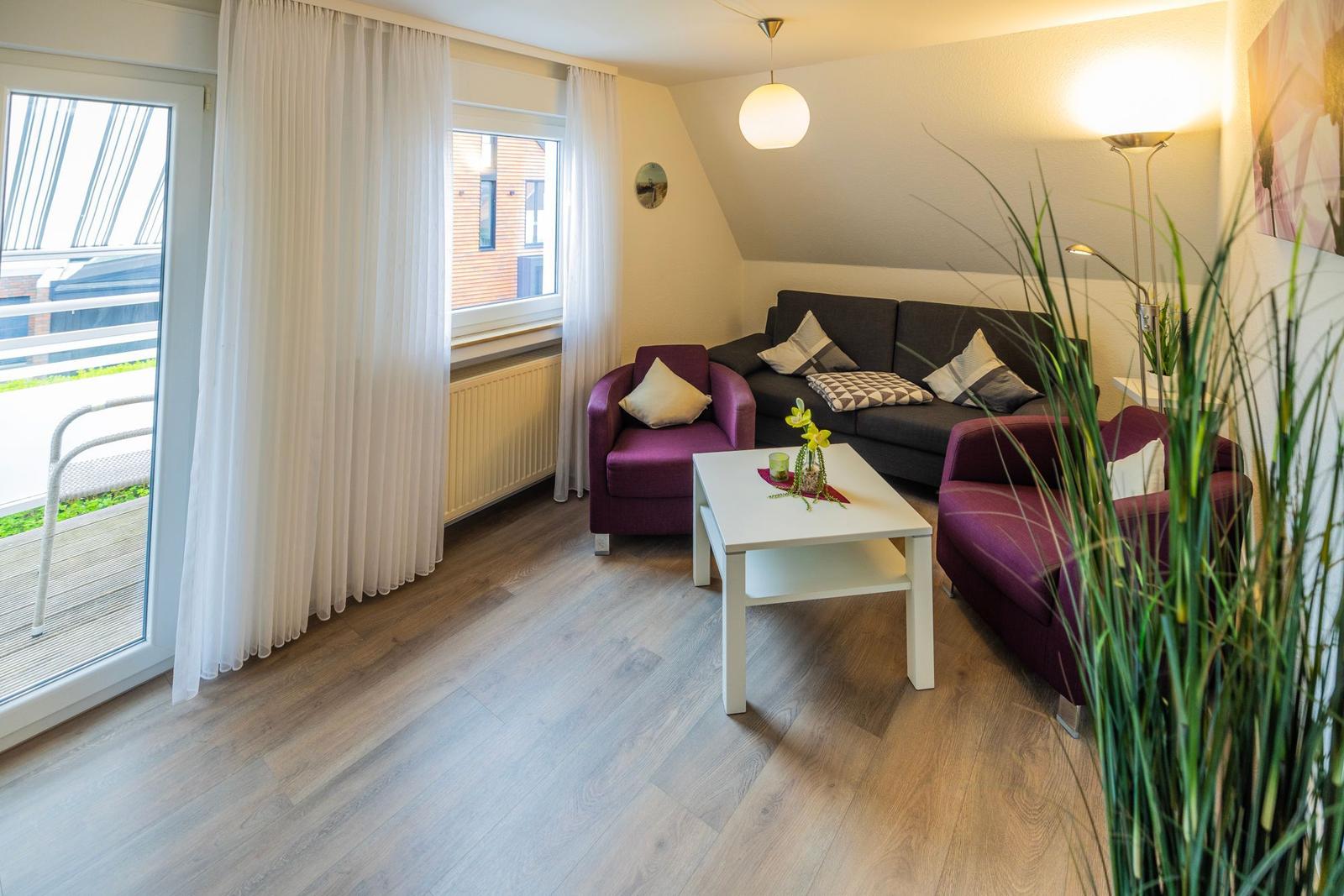 Wohnzimmer mit Sofa, zwei Sesseln und Tisch. Terrasse über Glastür. Holzboden und helle Wände.