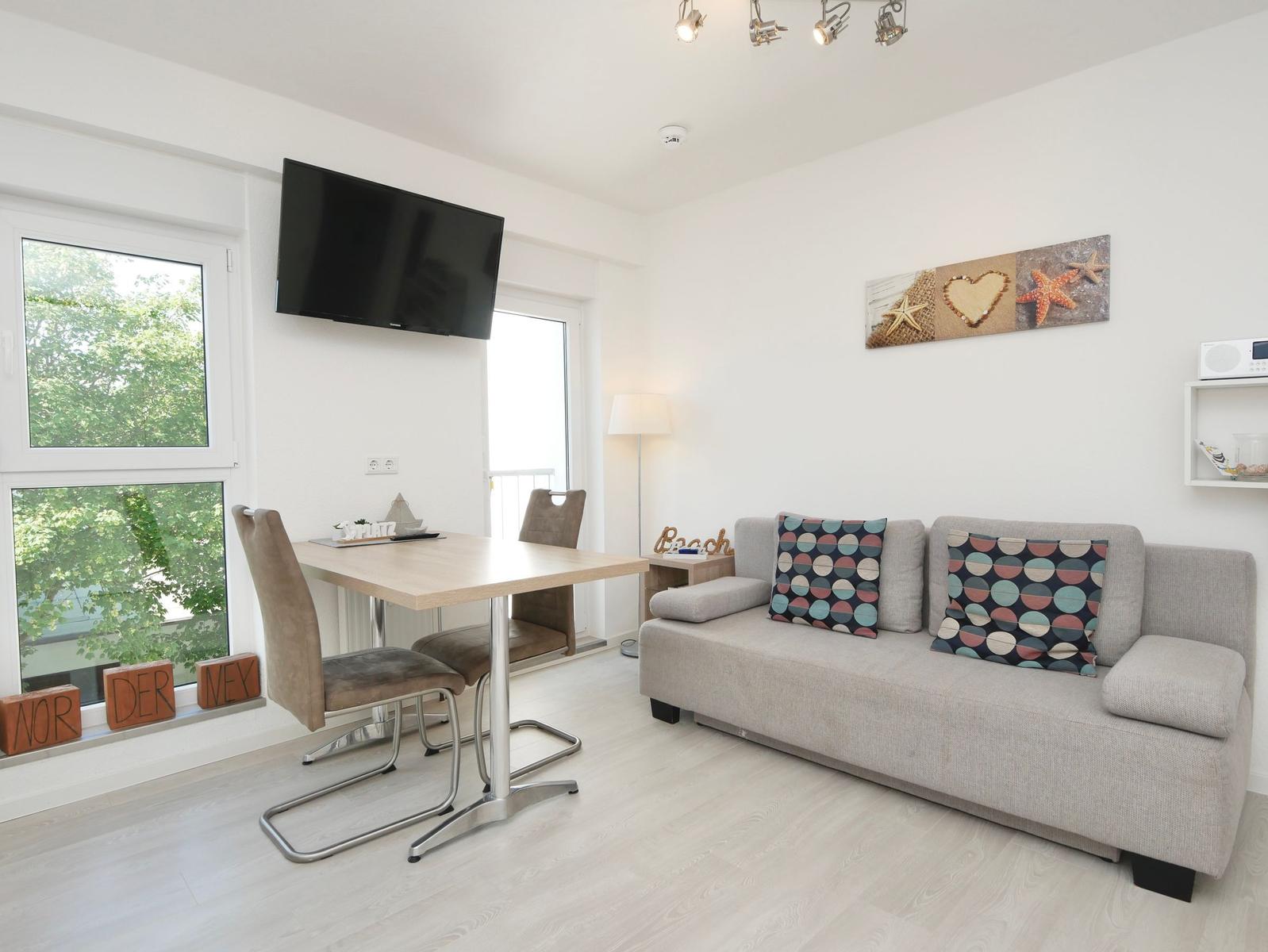 Wohnzimmer mit Sofa, Esstisch und Fernseher an der Wand. Fenster mit Blick auf Grün.