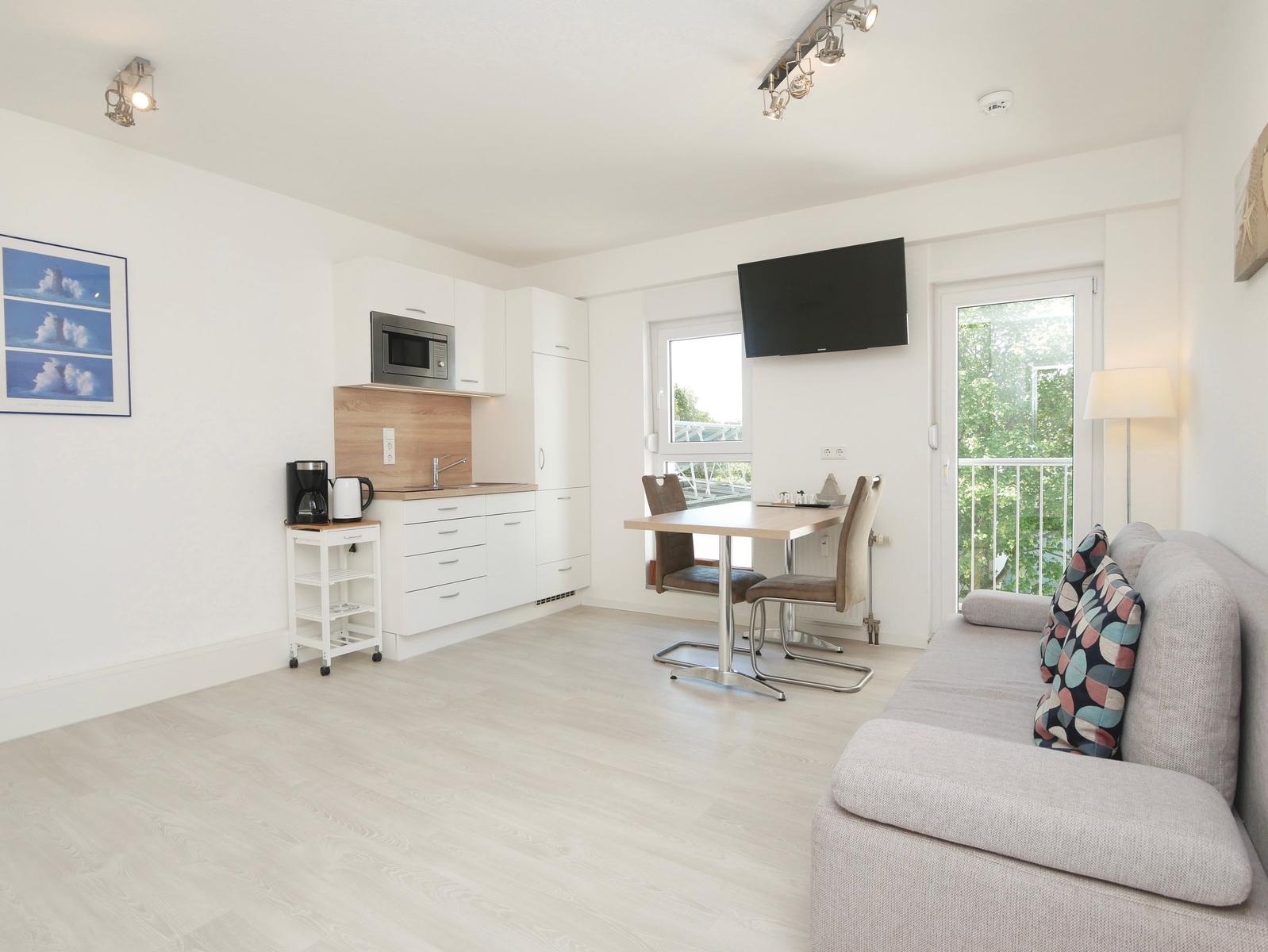 Wohnküche mit Sofa, Esstisch und Balkontür. Küche mit Mikrowelle und Kühlschrank.