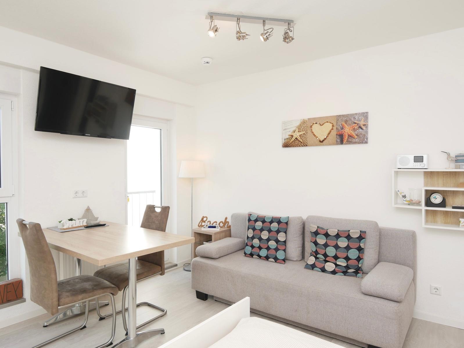 Wohnzimmer mit Sofa, Esstisch und Fernseher an der Wand.