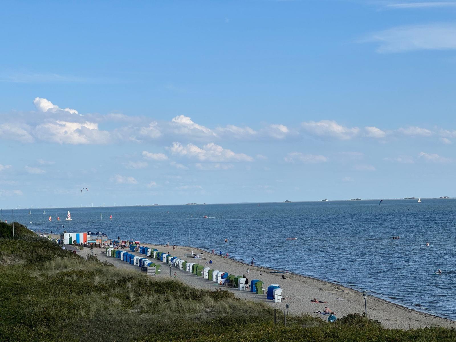 Strand mit bunten Strandkörben, Booten und Kitesurfern unter blauem Himmel.