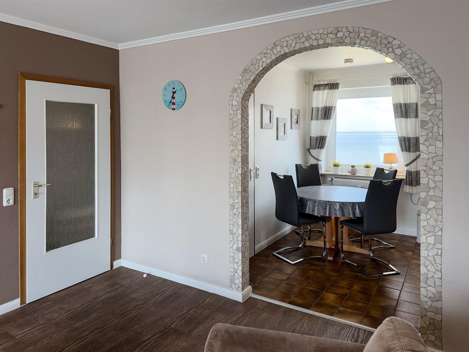 Through a stone arch, a dining area with table and chairs is visible. A clock hangs on the wall.