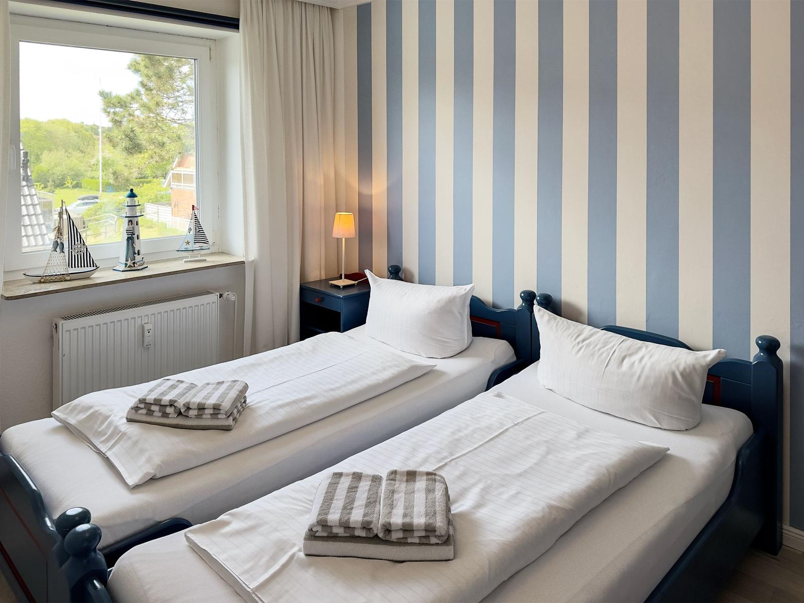 Two beds with white bedding and towels. Striped wall, window with view outside.