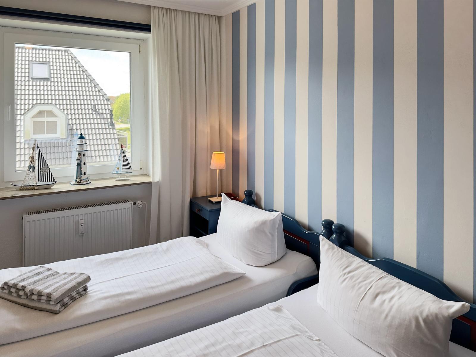 Two beds with white sheets, striped wall, and window with view of a house.