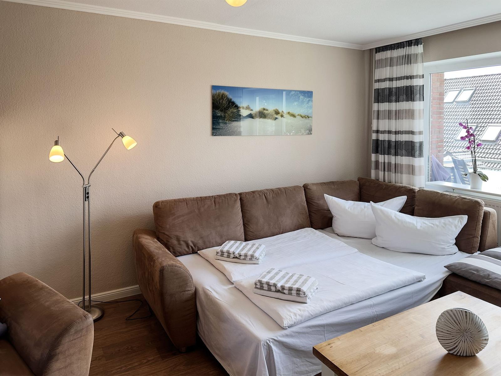 A bedroom with a brown sofa, white bedding, and a window overlooking rooftops.