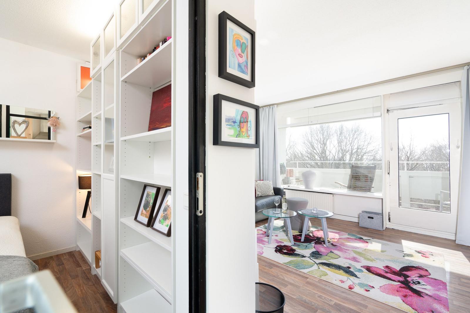 Living room with window, balcony, and floral rug. Bookcase and pictures on the wall.