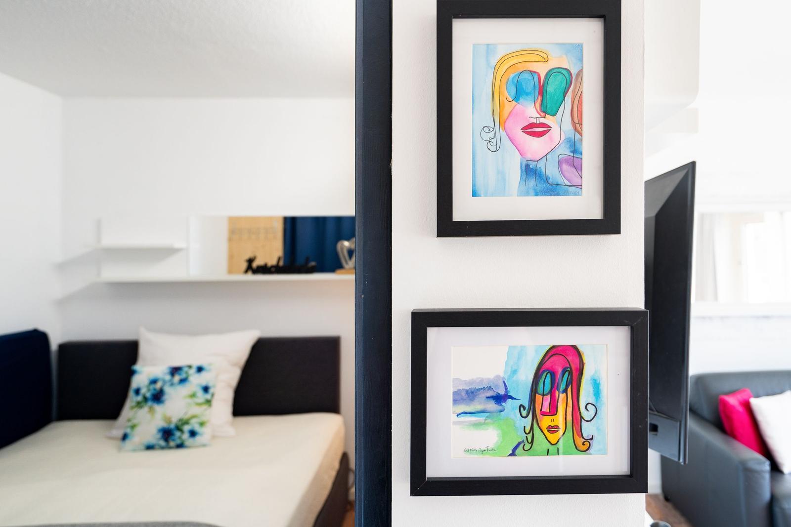 Two colorful paintings on the wall. In the background, a bed and a sofa are visible.