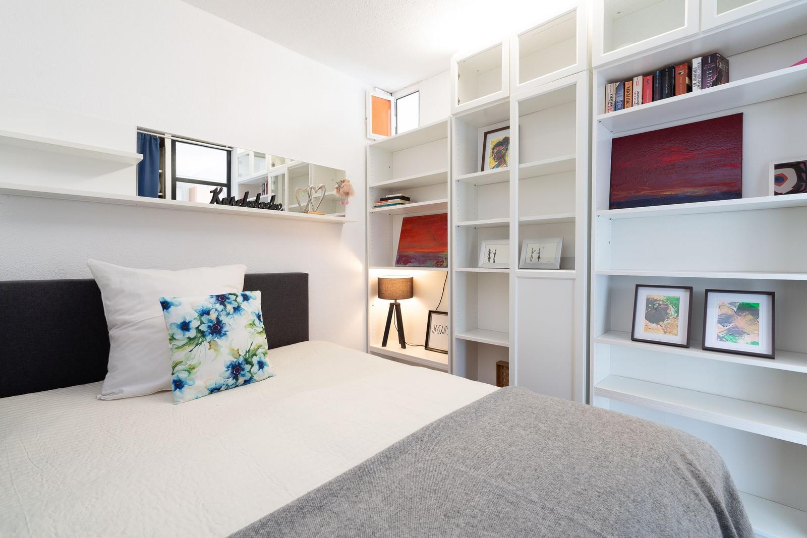 Bedroom with bed, white shelves, and window. Decorated with pictures and pillows.